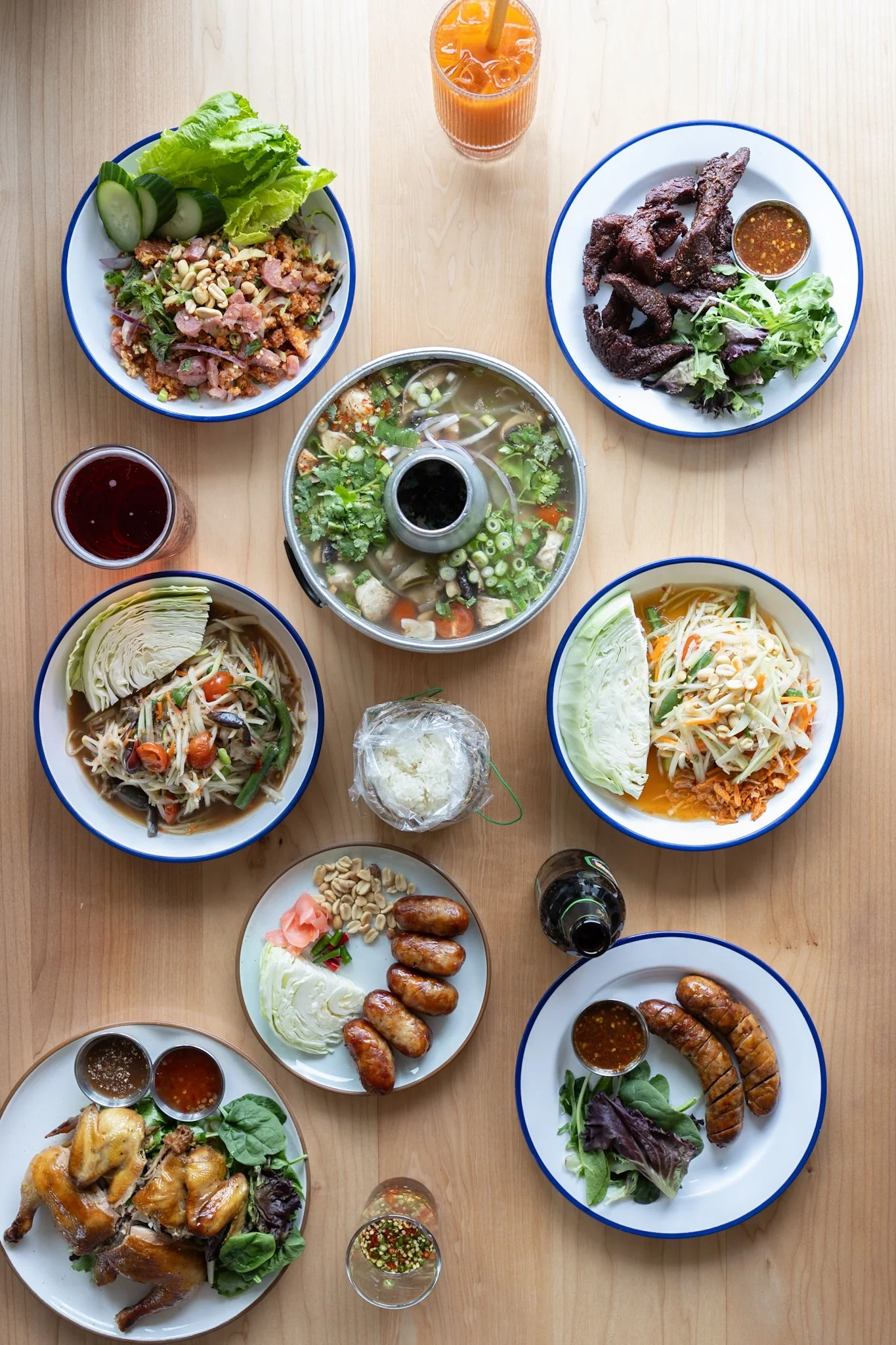 Various plates of Asian dishes including grilled meat, soups, salads, spring rolls, and dipping sauces on a wooden table with beverages.