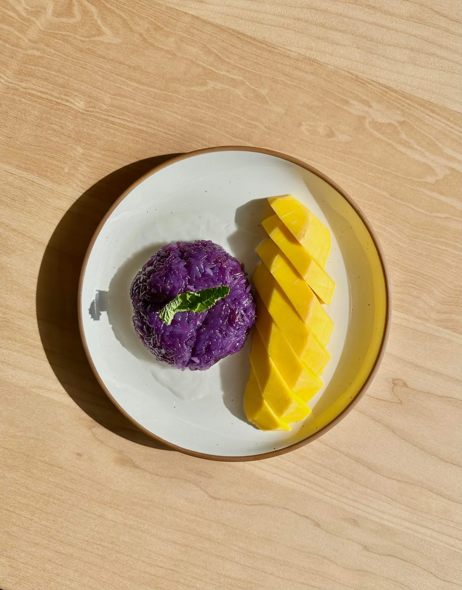 A plate with sliced mango and a purple dessert garnished with a mint leaf on a wooden surface.