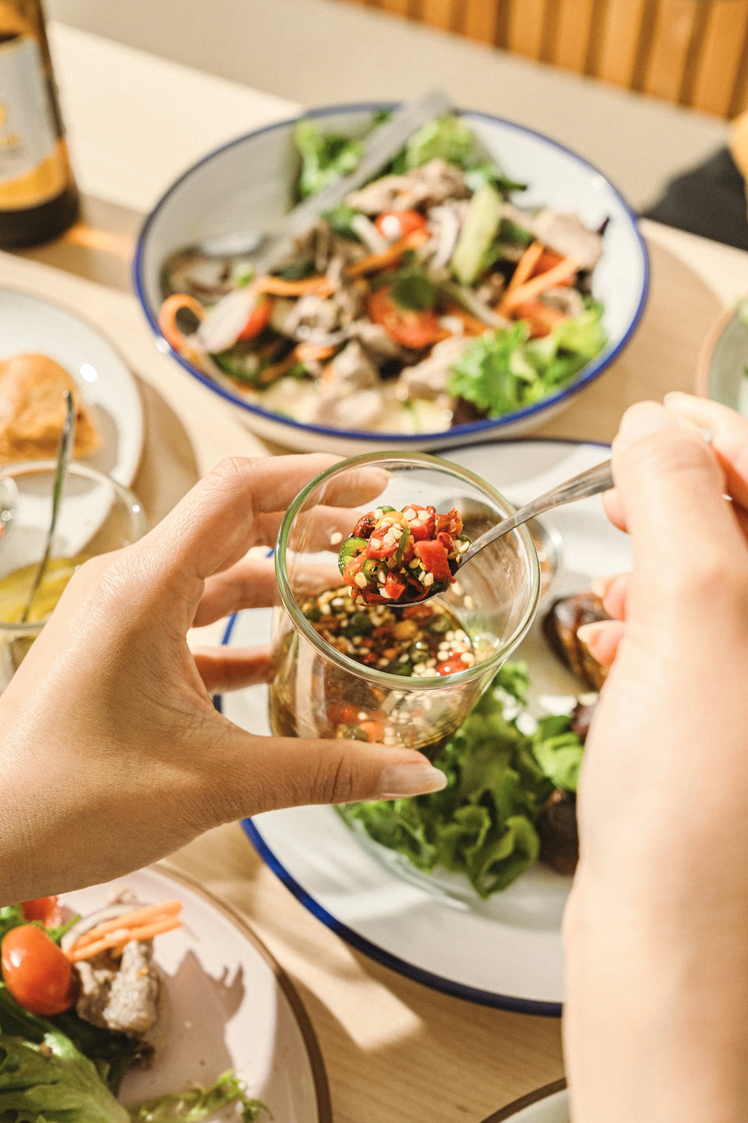 Close-up of hands holding a small glass jar with chopped red and green chili peppers, with a large bowl of a vegetable and meat salad in the background.