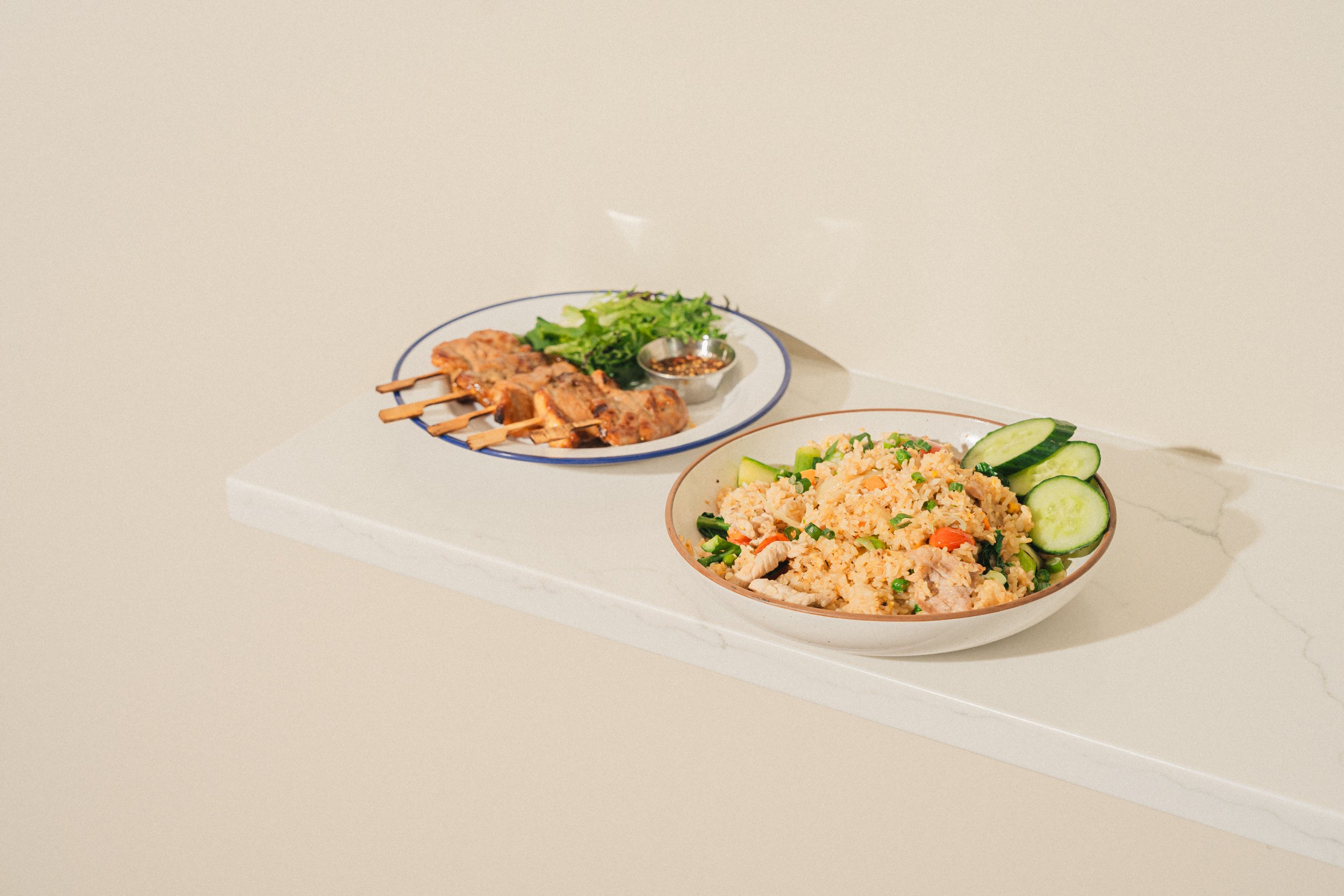 A white table with a bowl of fried rice garnished with cucumber slices, and a plate of grilled skewered meat with a side of greens and dipping sauce.