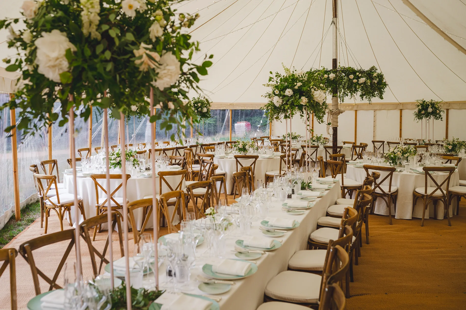 A traditional marquee with foliage decorations and white flowers. Chairs are rustic wood and table linens are all white.