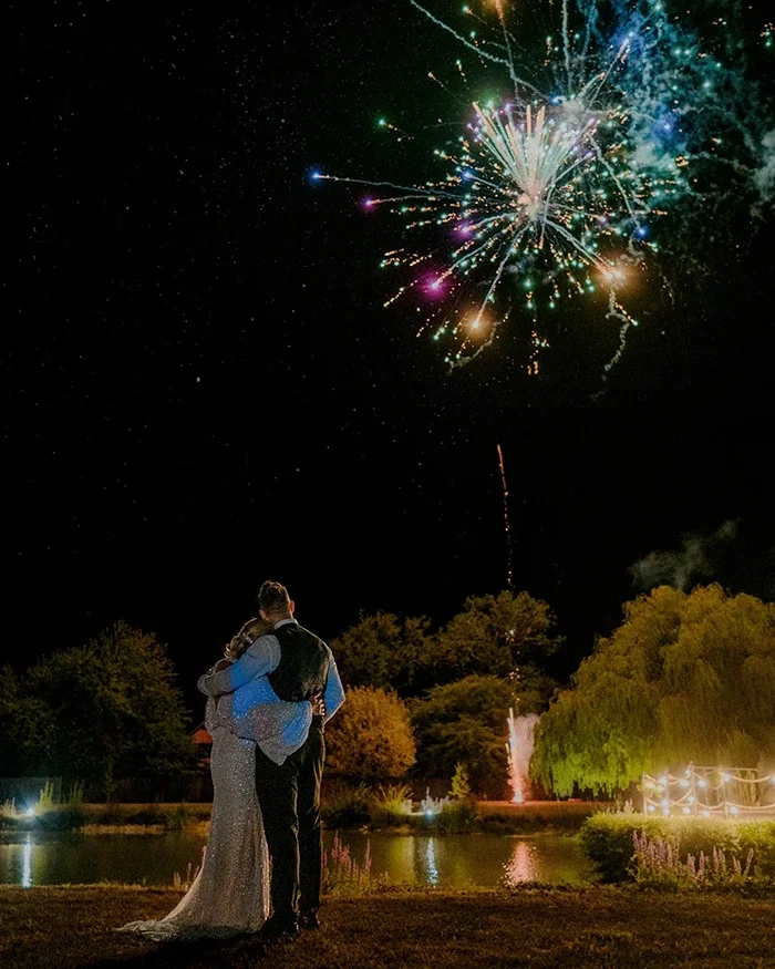 A newlywed couple watching fireworks at their evening wedding reception in Norfolk, overlooking a garden and pond