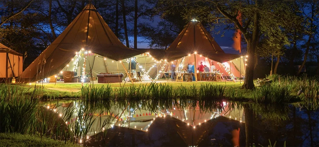 Tipis lit by festoon lights next to a reed-lined pond in the Norfolk Broads