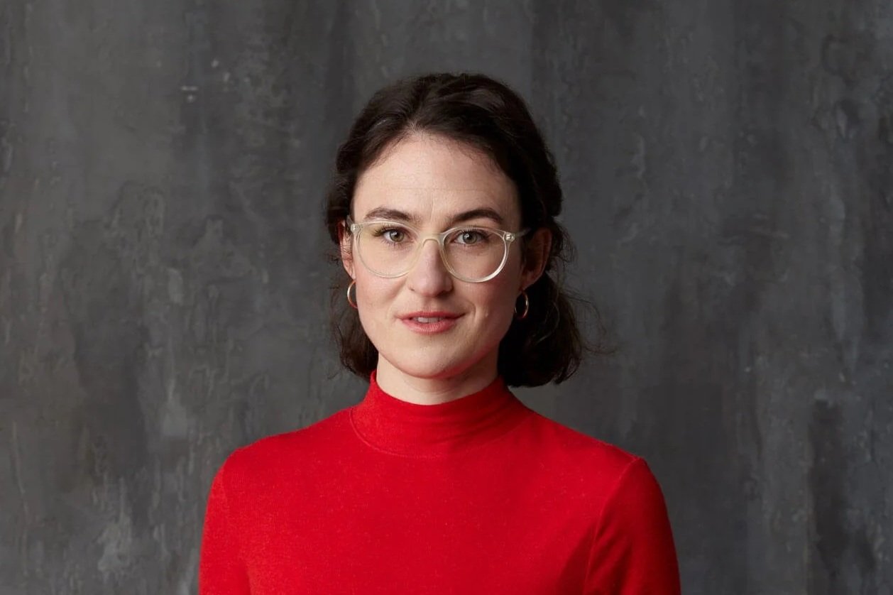 Eine junge Frau mit dunklen, lockigen Haaren, trägt eine rote Turtelneck und transparente Brille, vor einem grauen Hintergrund.