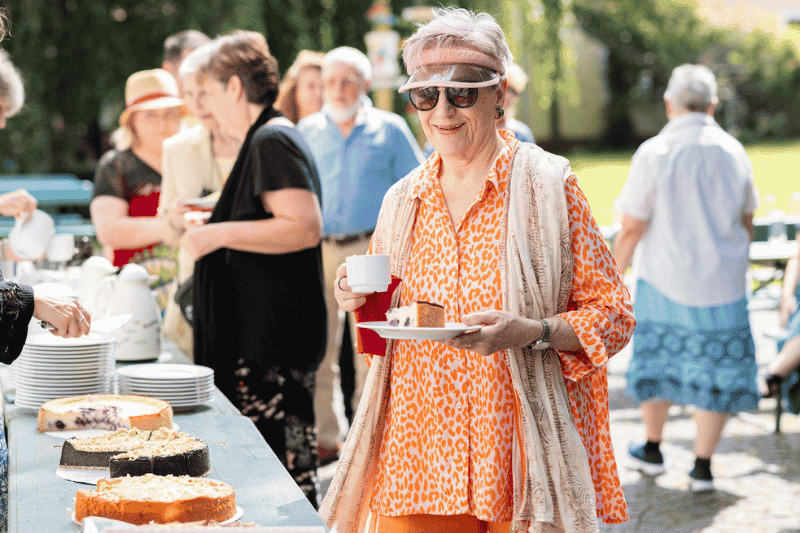 Ältere Frau mit Sonnenbrille und Sonnenvisier beim Kaffeetrinken bei einem Sommerveranstaltung von KulturRaum München, im Hintergrund mehr Menschen, die sich an einem Büffet mit Kuchen bedienen.