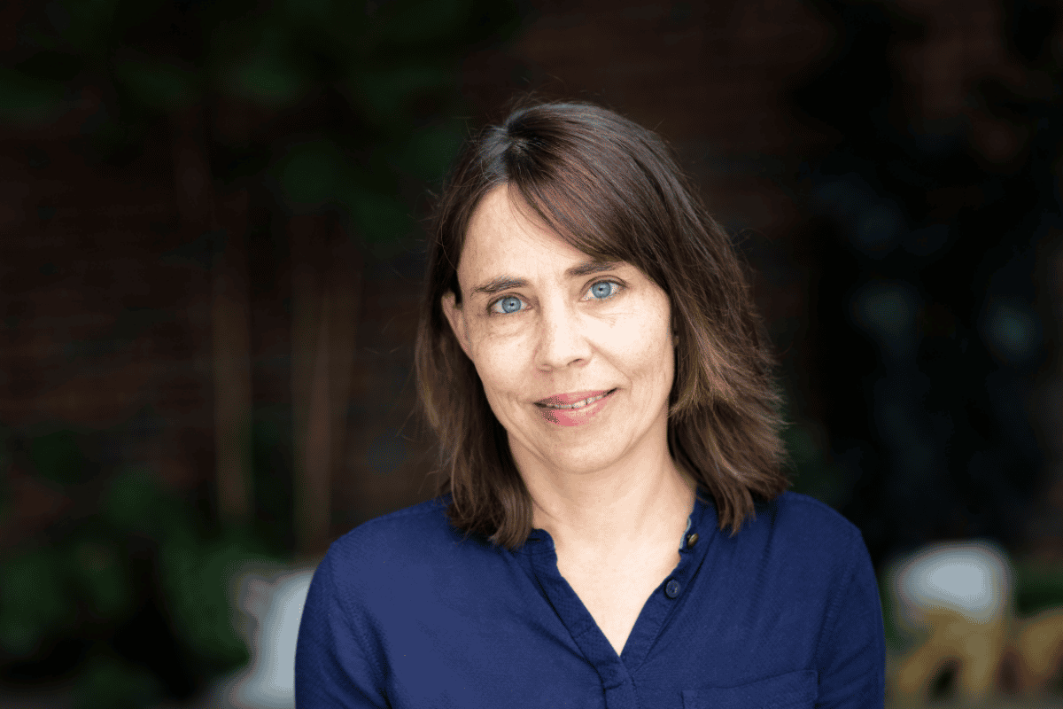 Portrait von Sabine Ruchlinski, die ein dunkelblaues Hemd trägt, vor einem unscharfen natürlichen Hintergrund.