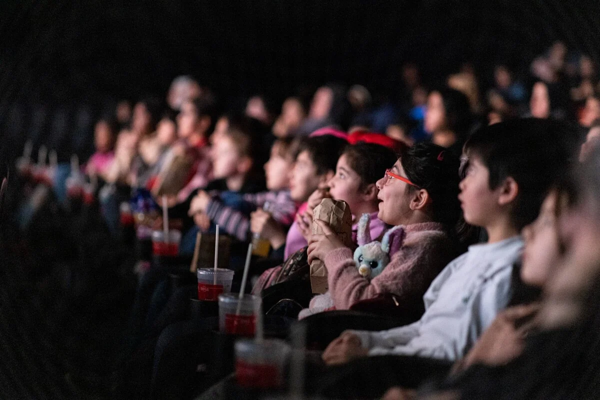 Kinder sitzen in einem dunklen Kino, schauen fasziniert einen Film und trinken Getränke.