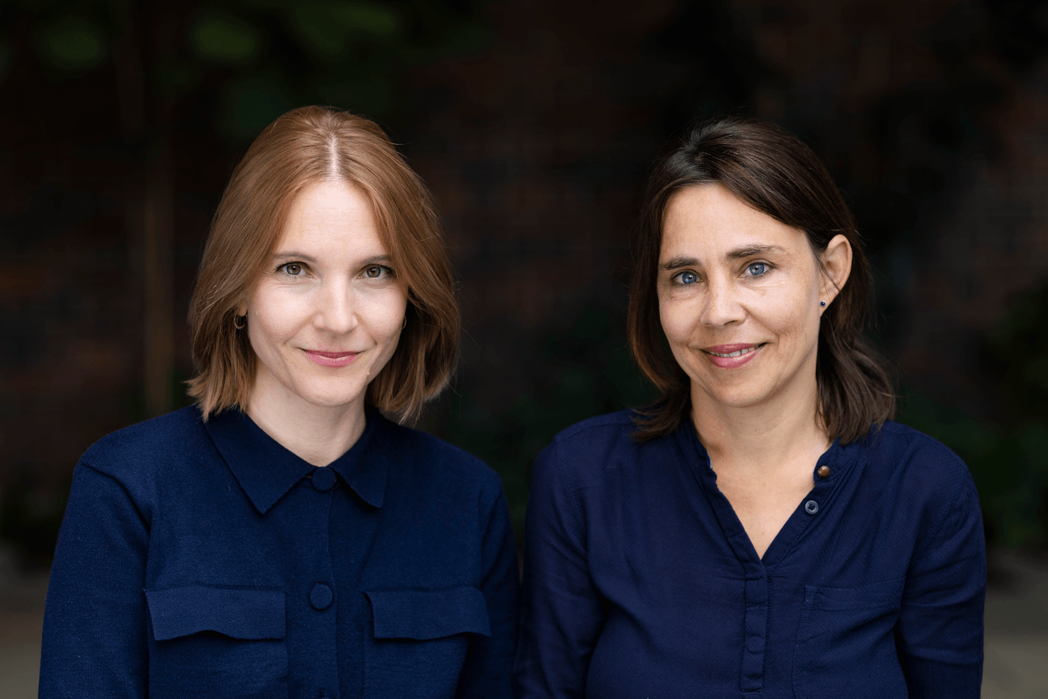 Katharina Maurer und Sabine Ruchlinski sitzen nebeneinander vor einem dunklen Hintergrund, beide tragen dunkelblaue Hemden und lächeln in die Kamera.