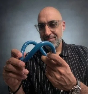 A bald man with glasses and a striped shirt holding a blue object with a loop and twist design.