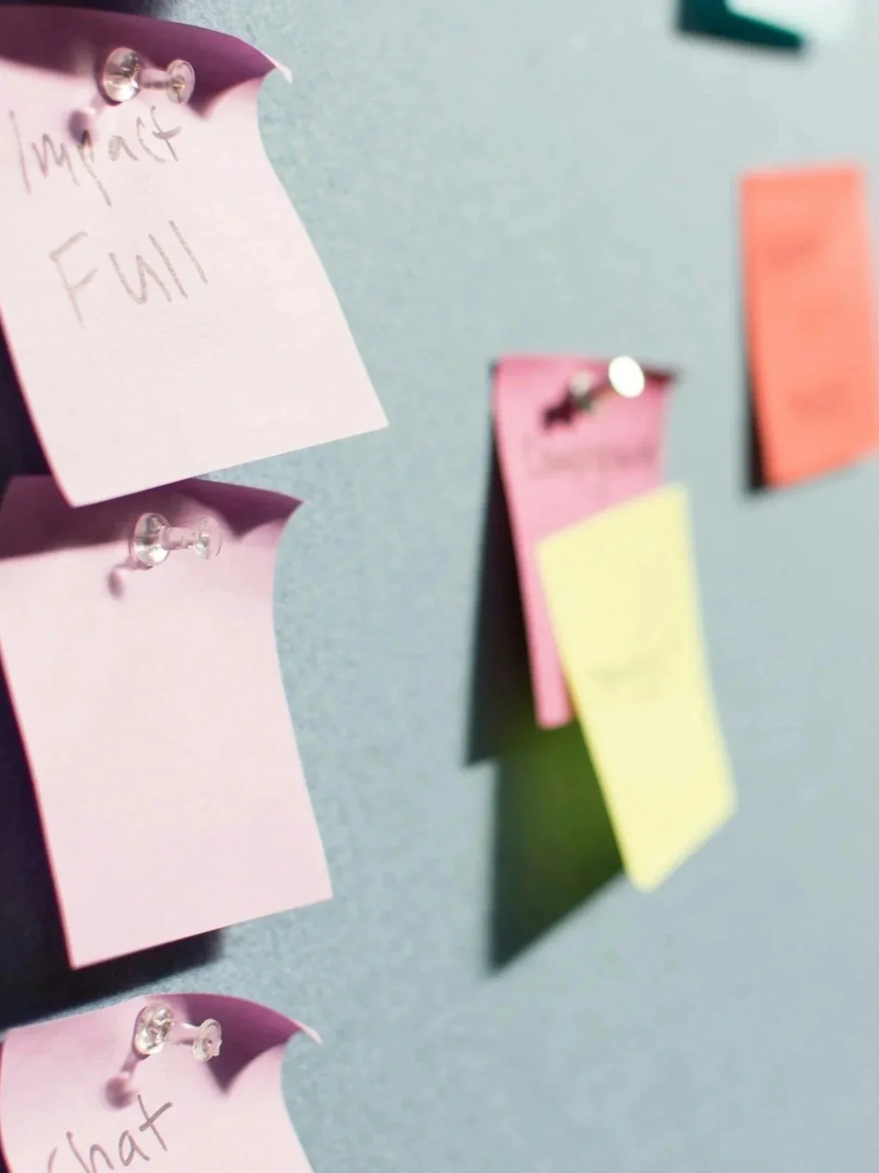 Close-up of a bulletin board with colorful sticky notes and a pink note with handwritten words, pinned with transparent push pins.