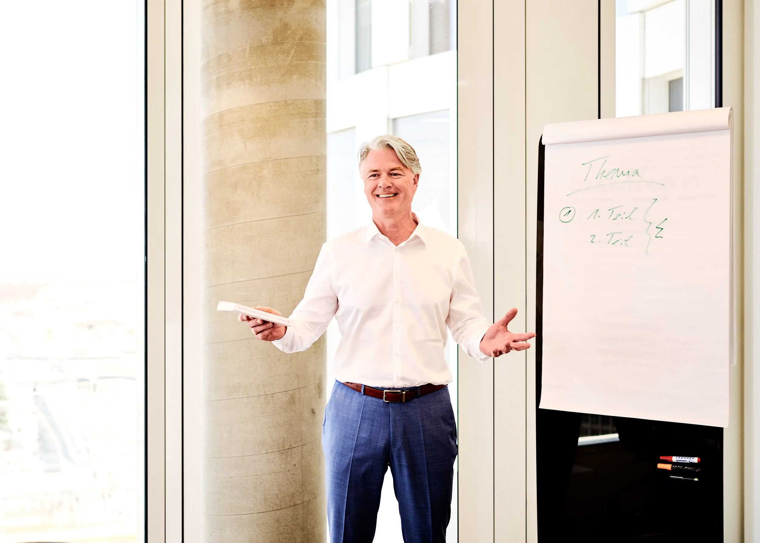 Ronny Weber von der Burg lächelt in einem weißen Hemd, der in einem modernen Raum vor einer Flipchart steht, die mit grüner Tinte beschriftet ist.