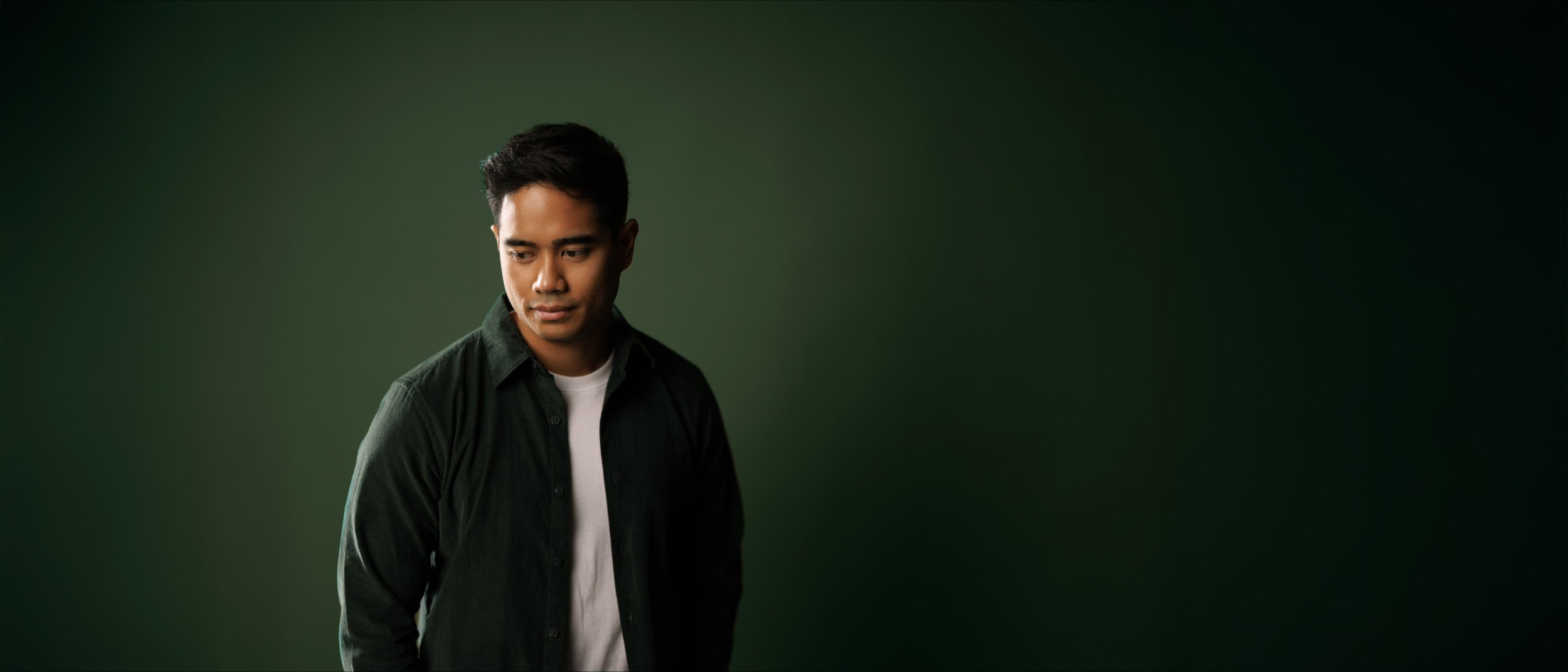 Portrait of composer Daniel O’Brien in a dark green shirt over a white T-shirt, looking down thoughtfully against a deep green backdrop