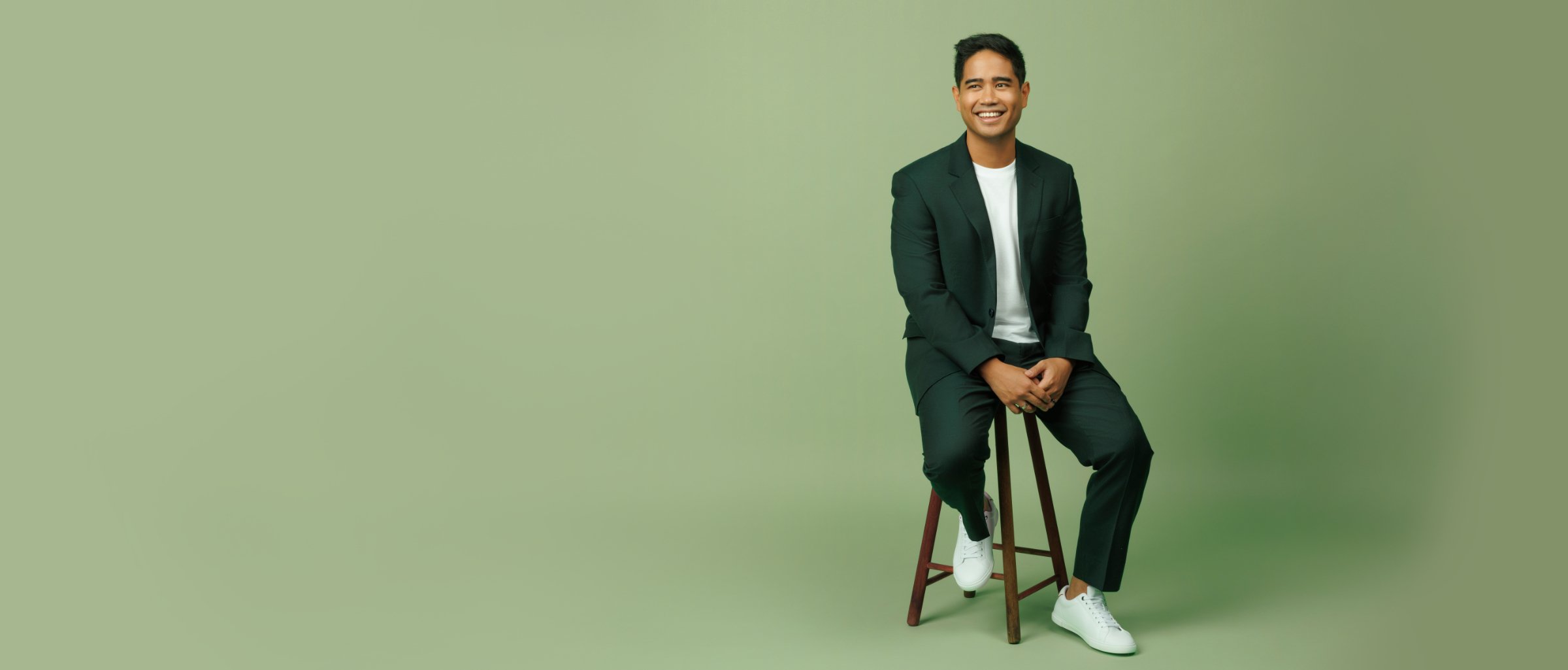 Composer Daniel O’Brien smiling, seated on a wooden stool in a dark green suit and white sneakers