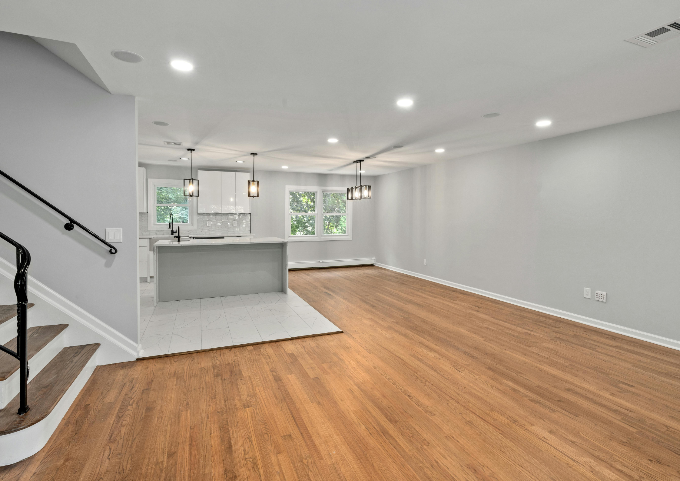 Empty living room with hardwood floors, white walls, ceiling with recessed lighting, a staircase on the left, and a kitchen area with pendant lights and windows in the background.