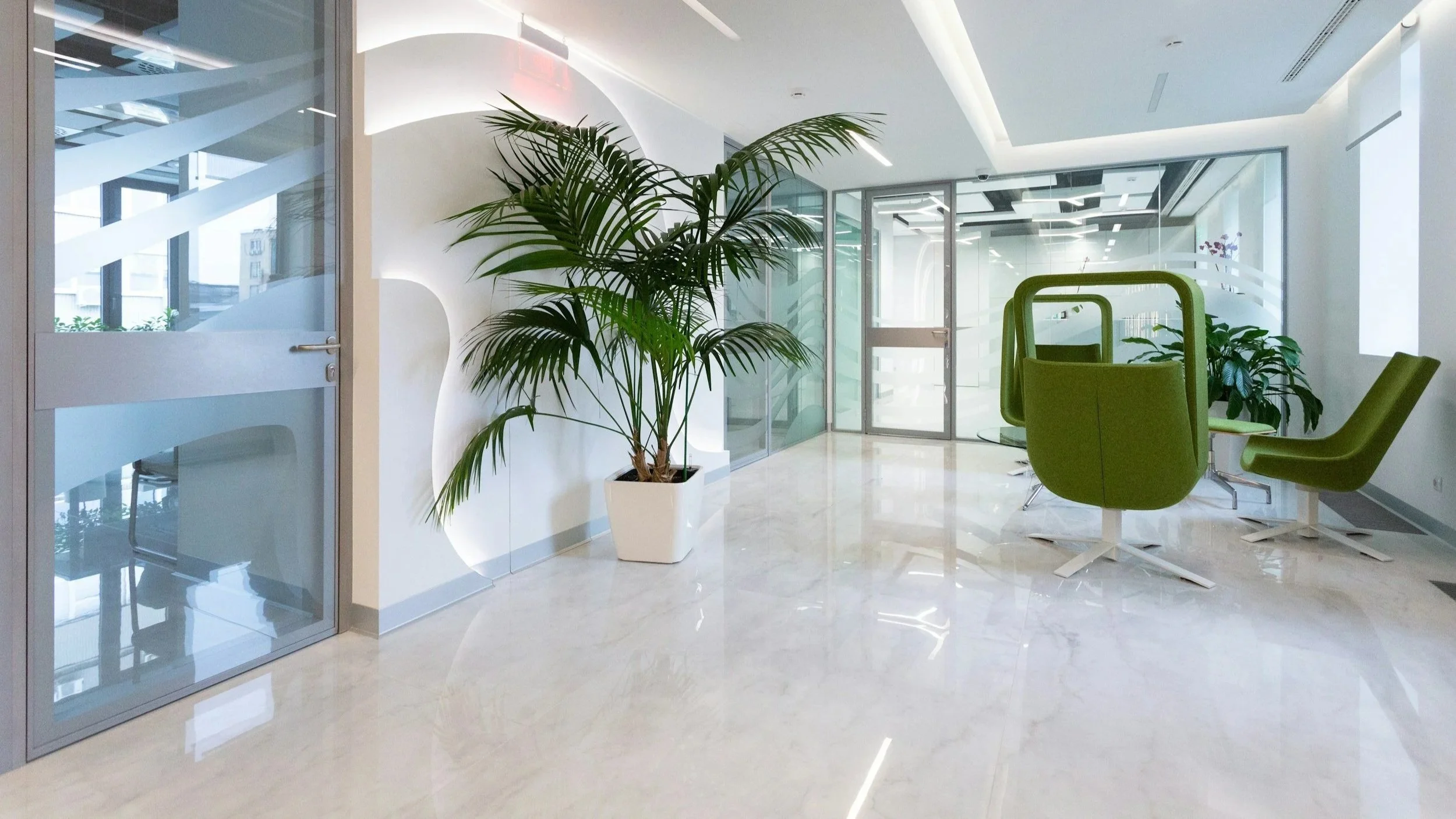 Modern office lounge area with green chairs, large potted plants, glass walls, and a marble floor.