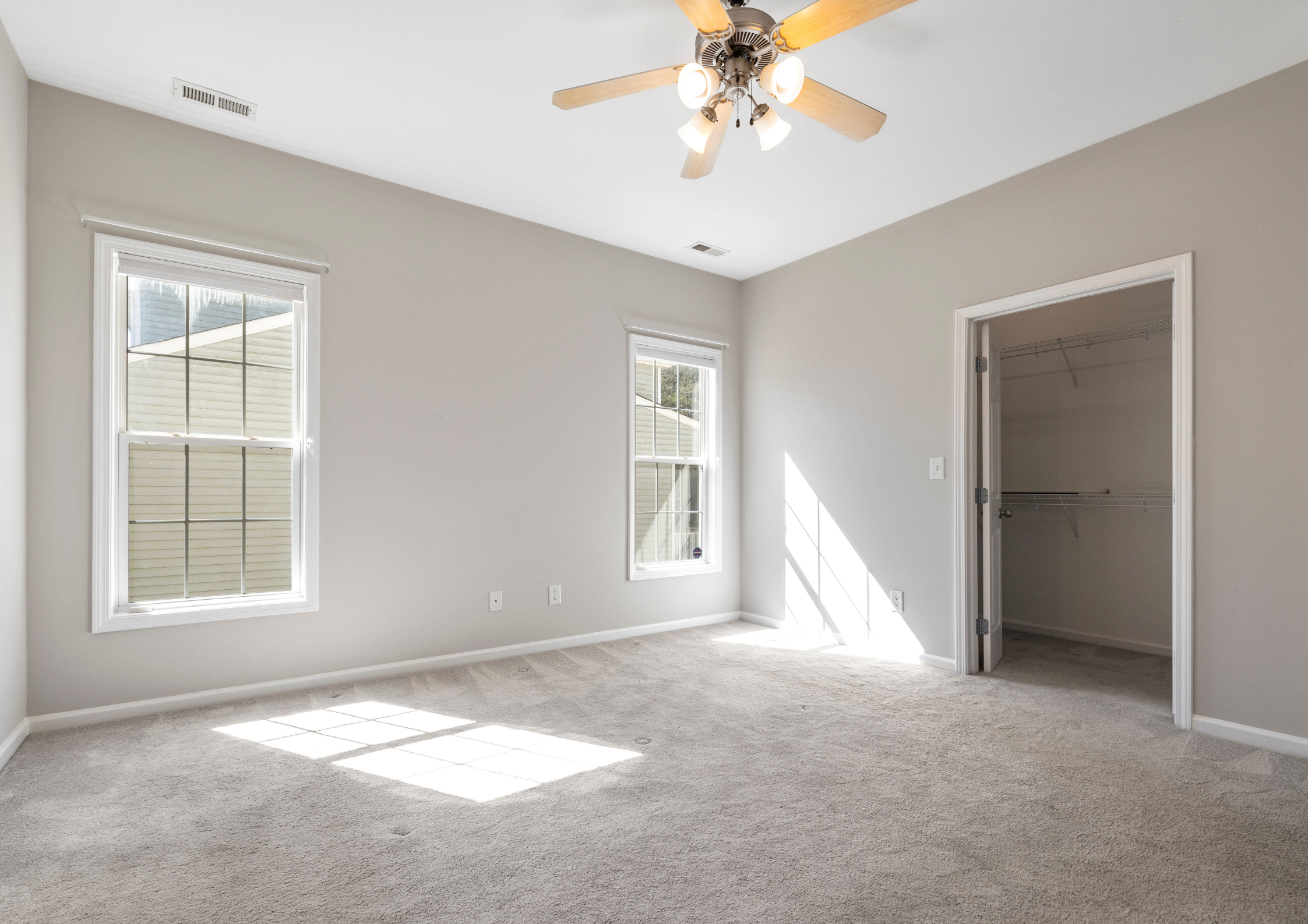 Empty room with beige carpet, two large windows, a ceiling fan with lights, and an open closet with wire shelves.