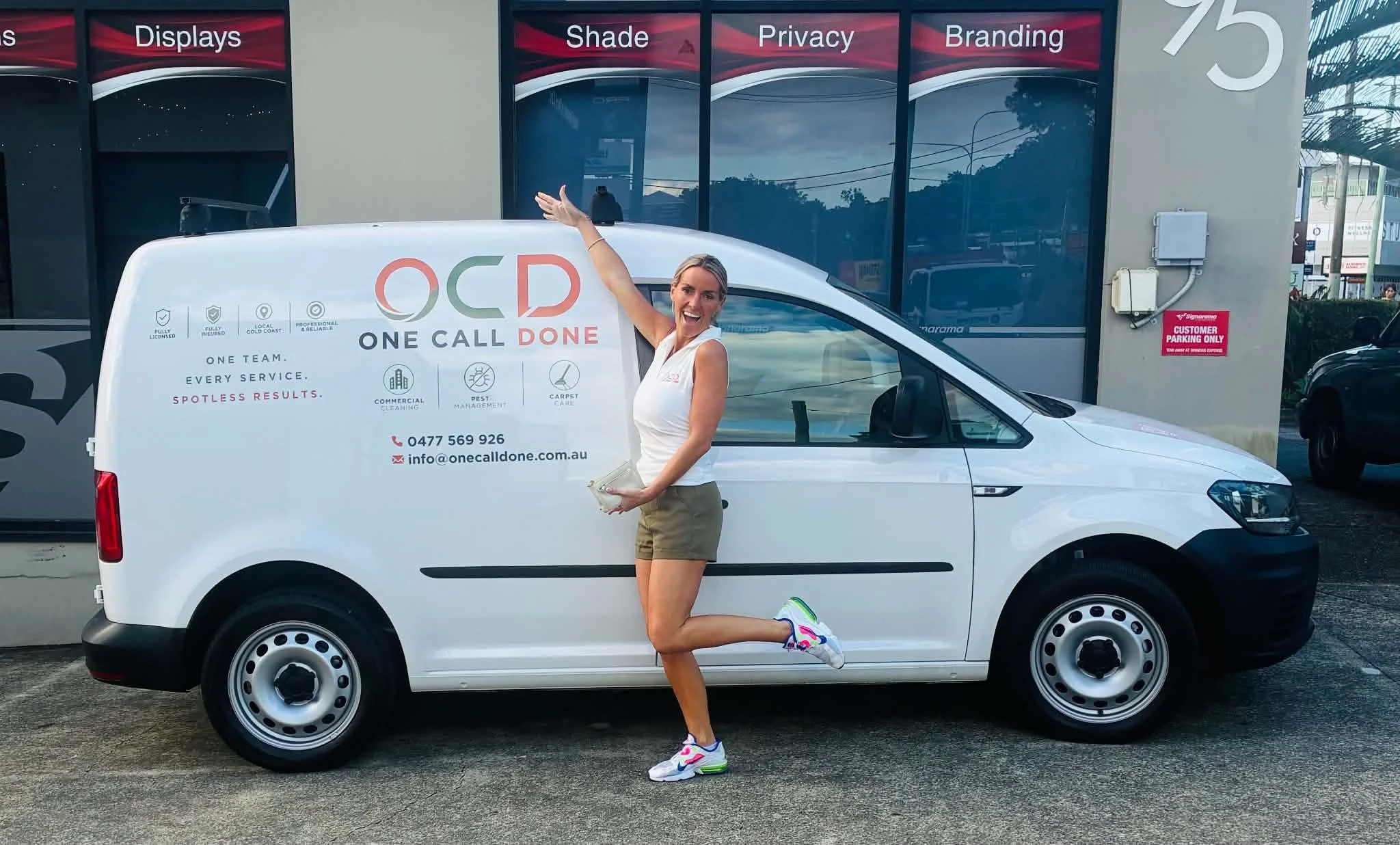 A woman with blonde hair, wearing a white sleeveless top, khaki shorts, and colorful running shoes, is smiling and posing with one knee bent on a white service van. The van has the logo 'OCD' and the words 'One Call Done' with icons and contact information on its side. In the background, there is a building with large window displays labeled 'Displays,' 'Shade,' 'Privacy,' and 'Branding.'