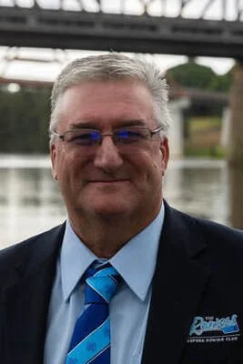 A middle-aged man with gray hair and glasses, dressed in a dark suit with a blue dress shirt and a striped blue tie, standing outdoors near a body of water with a bridge in the background.