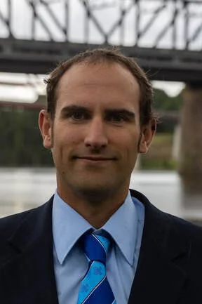 A man in a suit and blue tie standing outdoors with a bridge in the background.