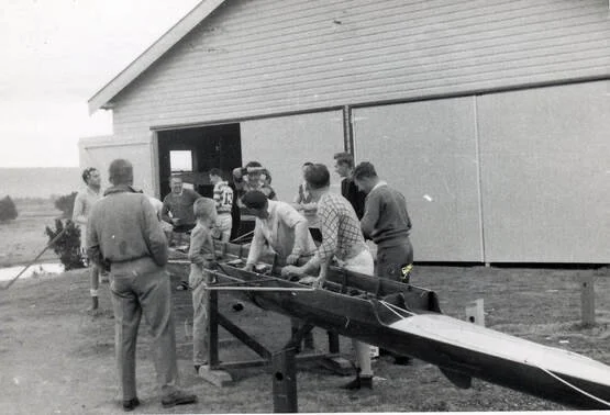 Group of people gathered around a boat outside a large building, some working on the boat while others observe.