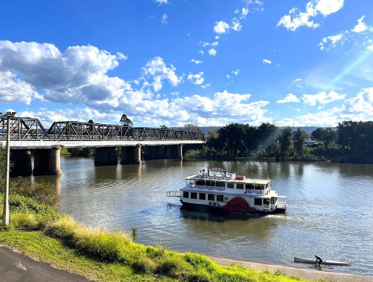 A boat sailing on a river with a tow bridge overhead and a paddleboarder nearby. The sky is partly cloudy with sun shining.