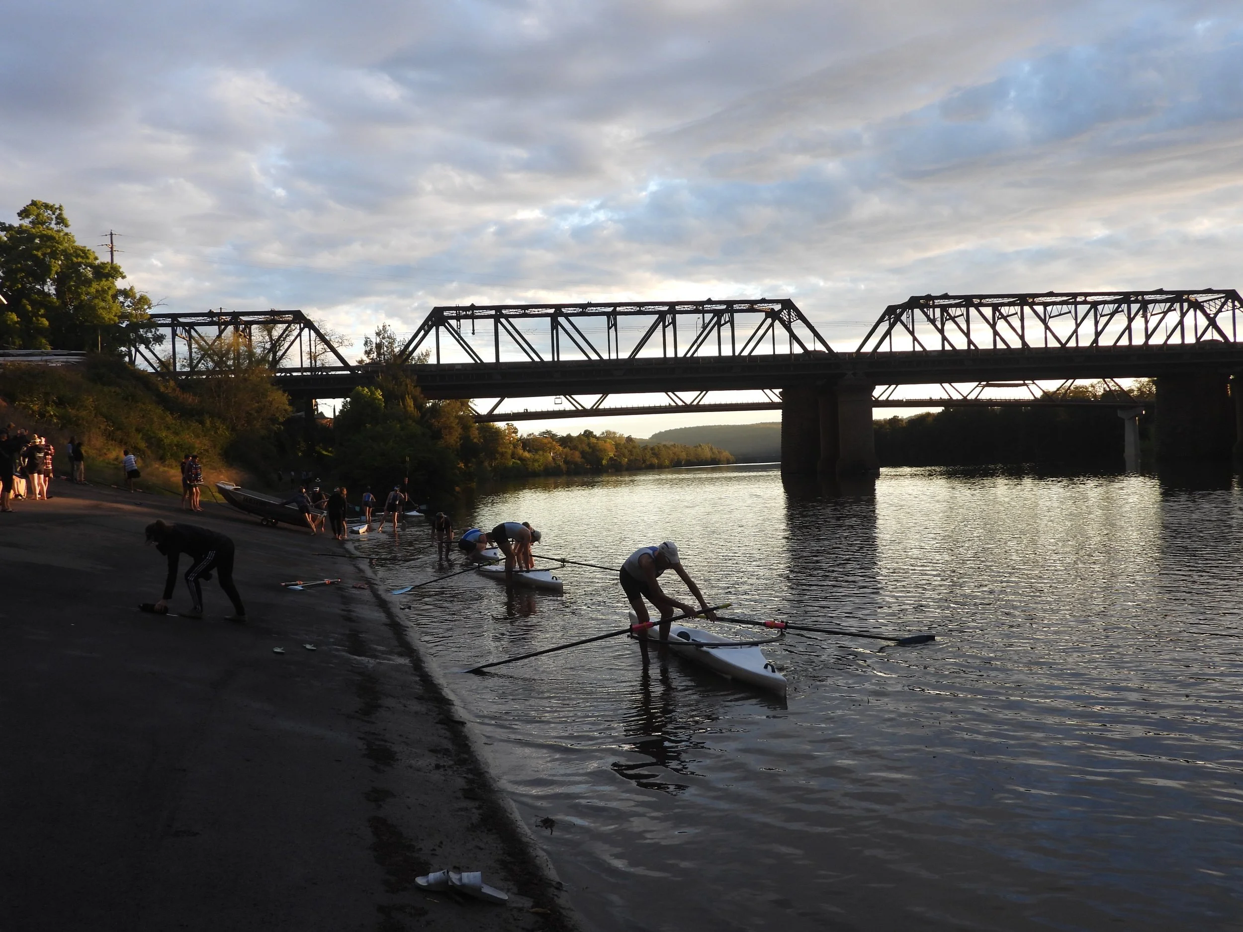 People preparing to launch sailboats into a river under a cloudy sky with a bridge overhead.