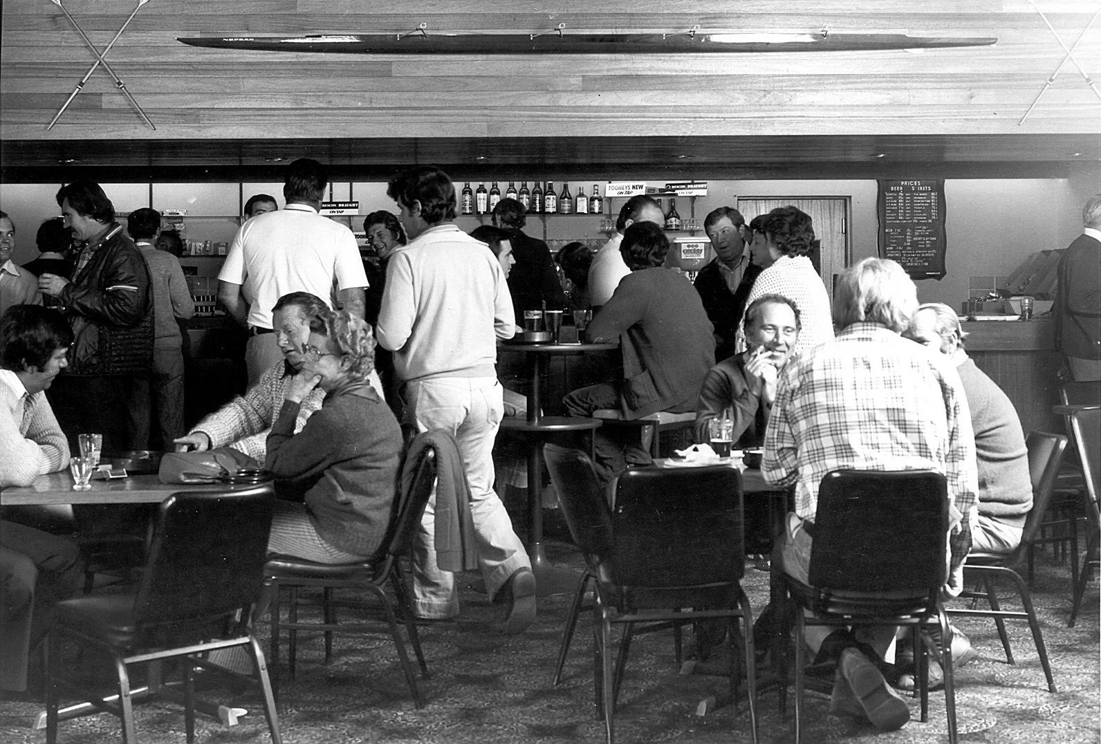 A black-and-white photograph of a crowded bar or restaurant with people sitting at tables and standing. Some patrons are engaged in conversations, and a bartender is behind the bar. The setting appears to be from the mid-20th century.