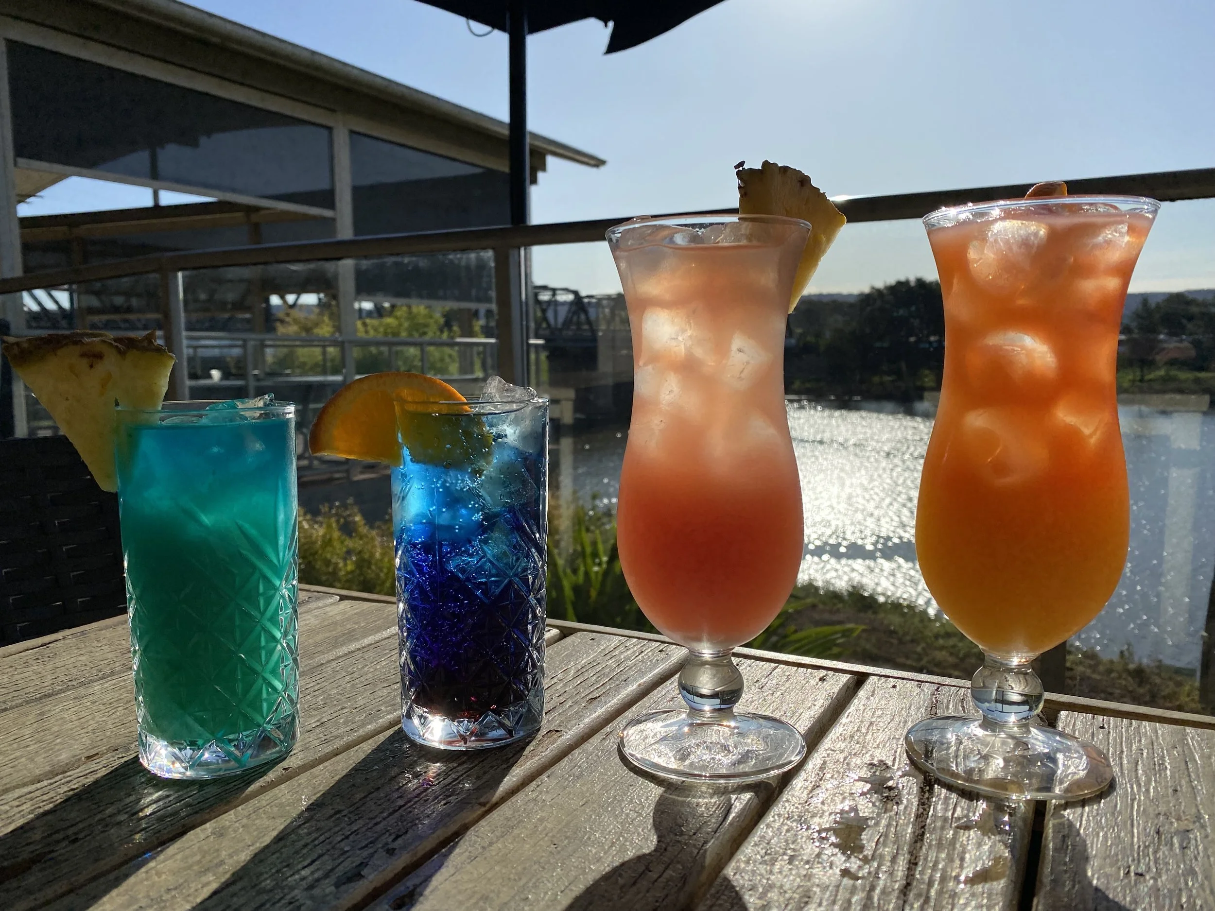 Four colorful cocktails on a wooden table with a scenic water view in the background. The drinks are garnished with pineapple and orange slices, and ice is visible in each glass.