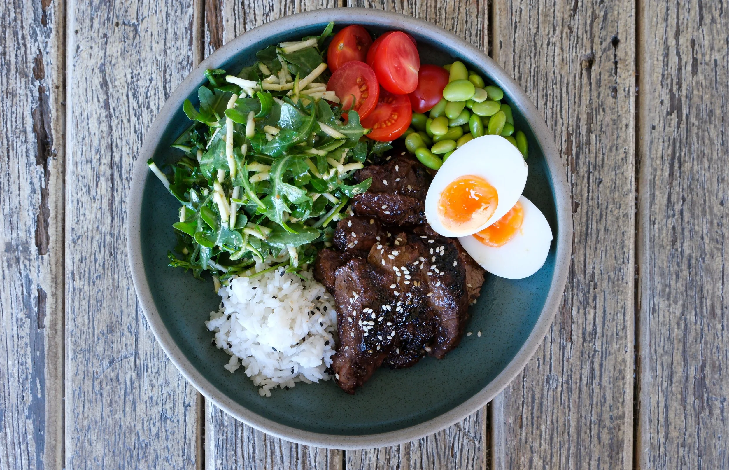 A bowl containing sliced cherry tomatoes, sliced hard-boiled eggs, cooked beef with sesame seeds, white rice, and a green salad with shredded cheese on top, placed on a weathered wooden surface.