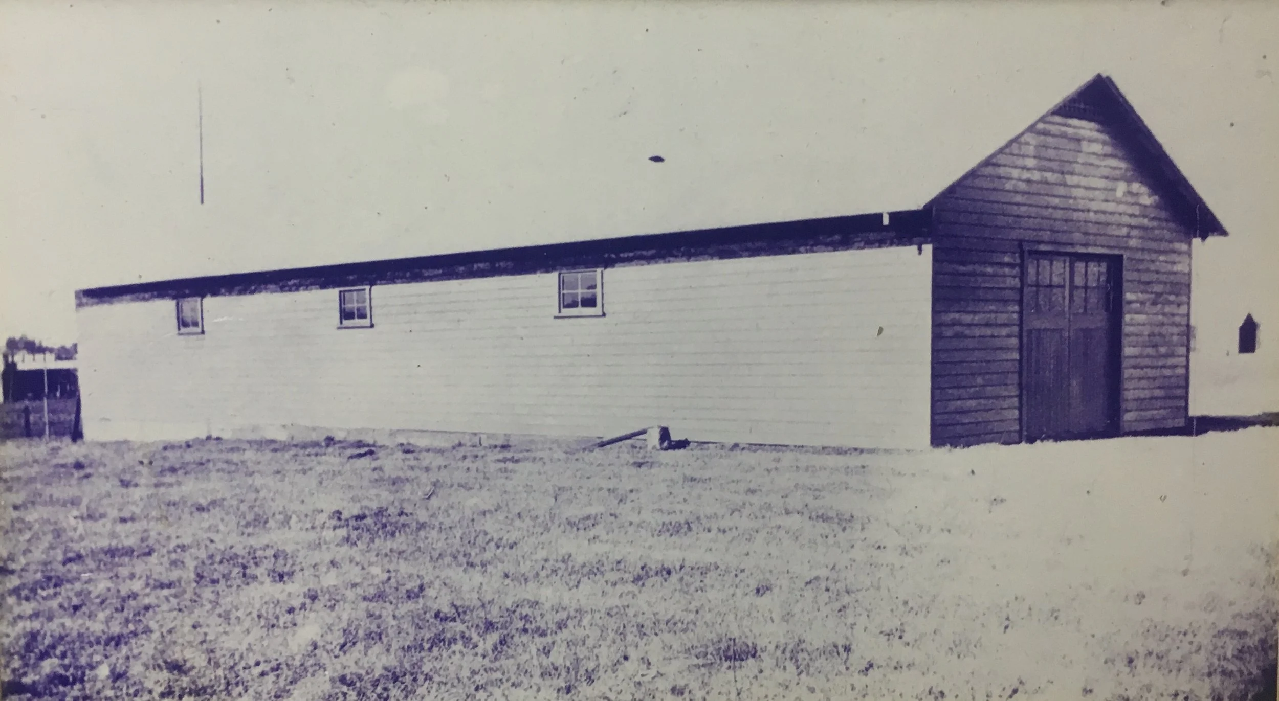 A black-and-white photo of a long, wooden barn or shed with a gable roof and four small windows along the side. The barn’s right end has a taller, square section with a large door. The ground in front is grassy, and there are a few small objects or structures in the background to the left.