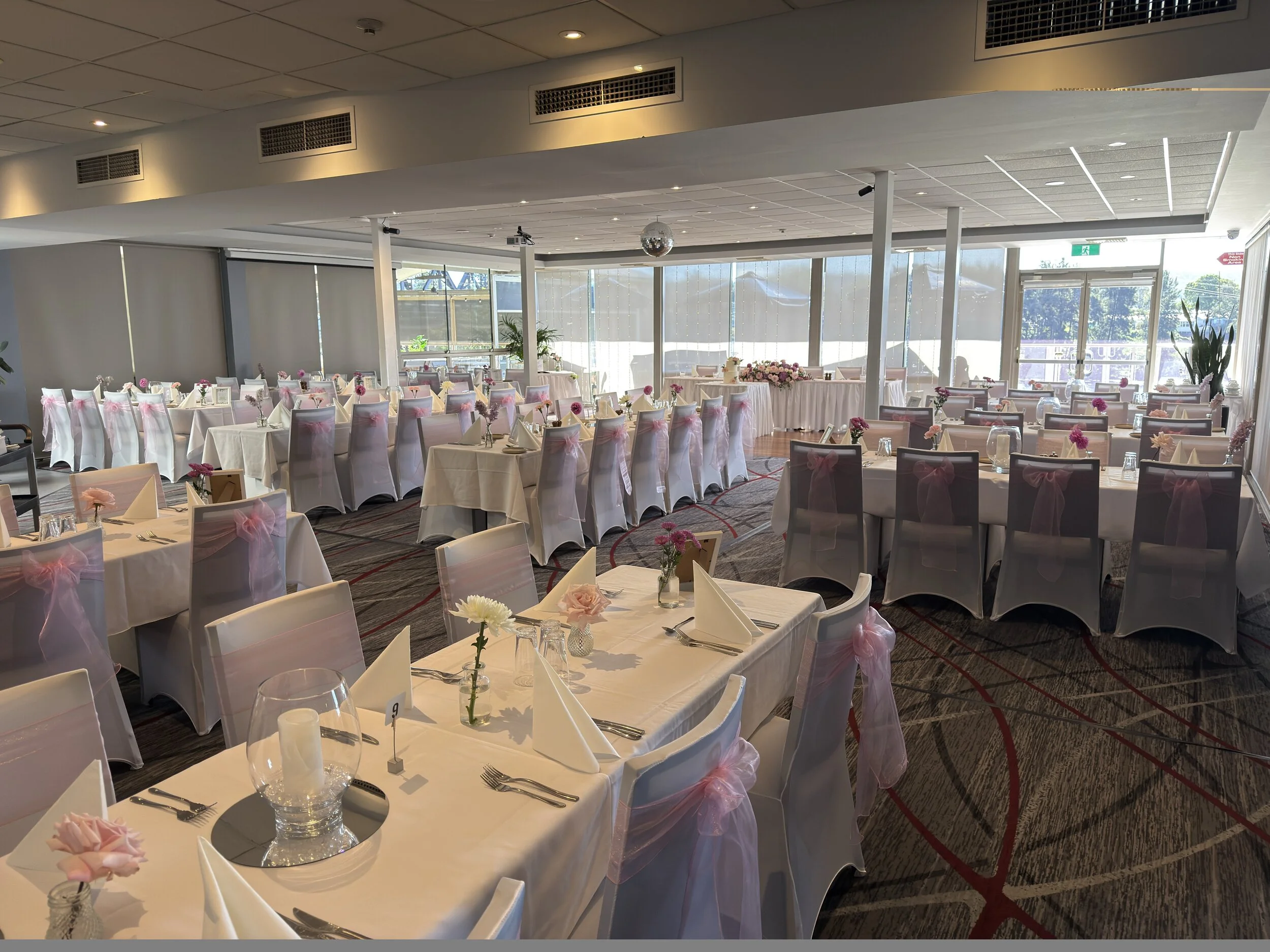 Dining hall or event space decorated with white tablecloths, pink ribbons, and floral centerpieces, set up for a celebration or formal gathering, with large windows and natural lighting.