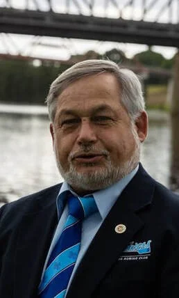A middle-aged man with gray hair and a beard, wearing a dark suit and a blue striped tie, standing outdoors near a body of water with a bridge structure in the background.