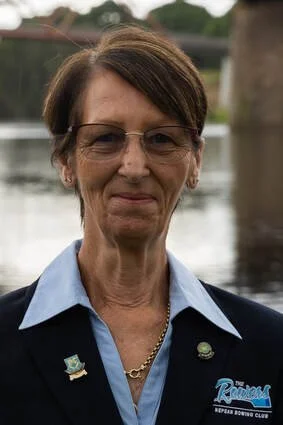 A woman with short brown hair, glasses, wearing a dark blazer with pins, a light blue collared shirt, and a gold necklace, standing outdoors near water.
