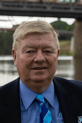 A middle-aged man in a suit and tie standing outdoors near water with a bridge in the background.