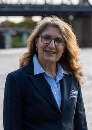 A woman with wavy, shoulder-length hair and glasses, wearing a dark blazer over a light blue collared shirt, standing outdoors near a river with a bridge in the background.