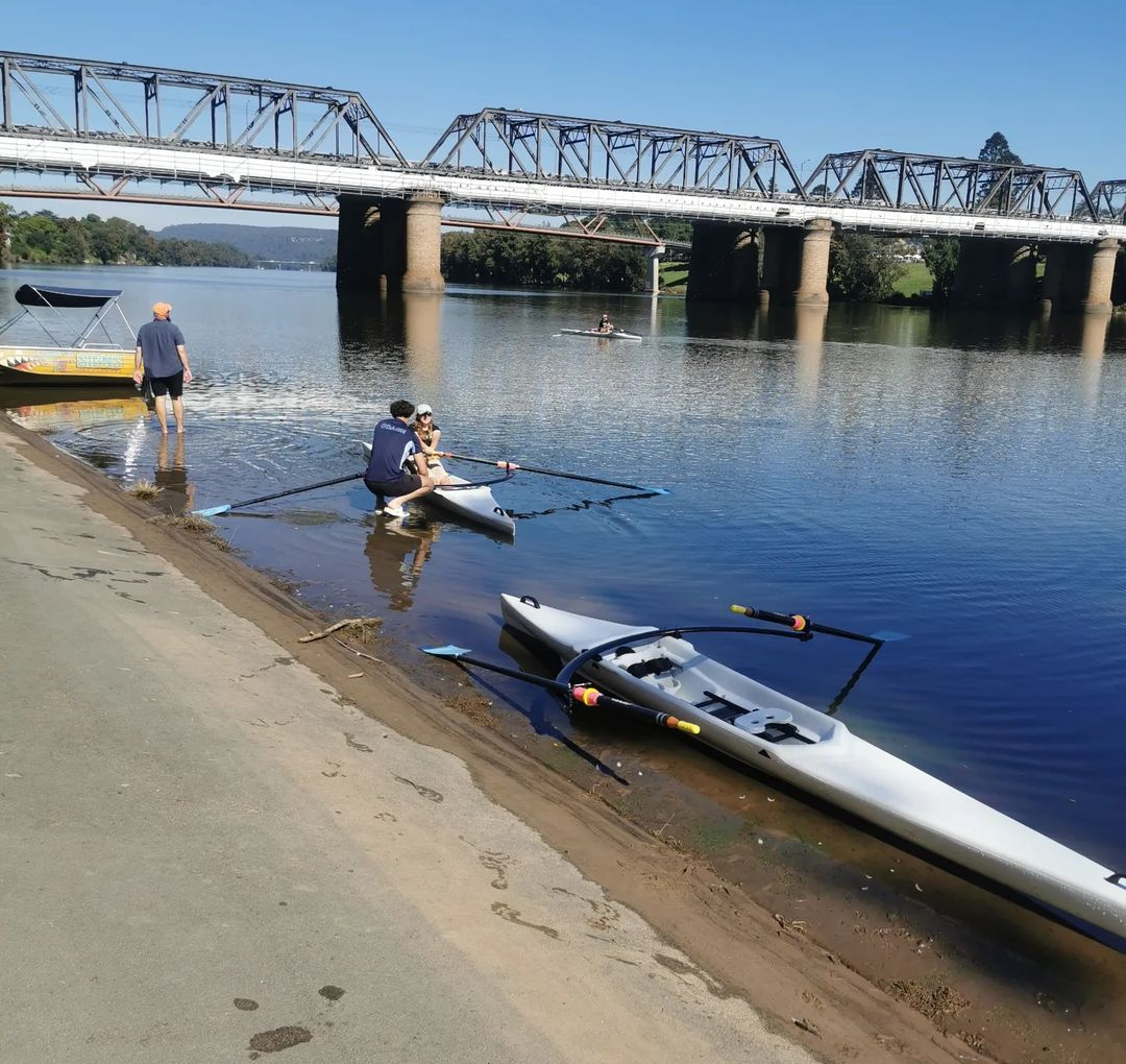 People launching and preparing kayaks near a riverbank with a train bridge overhead.