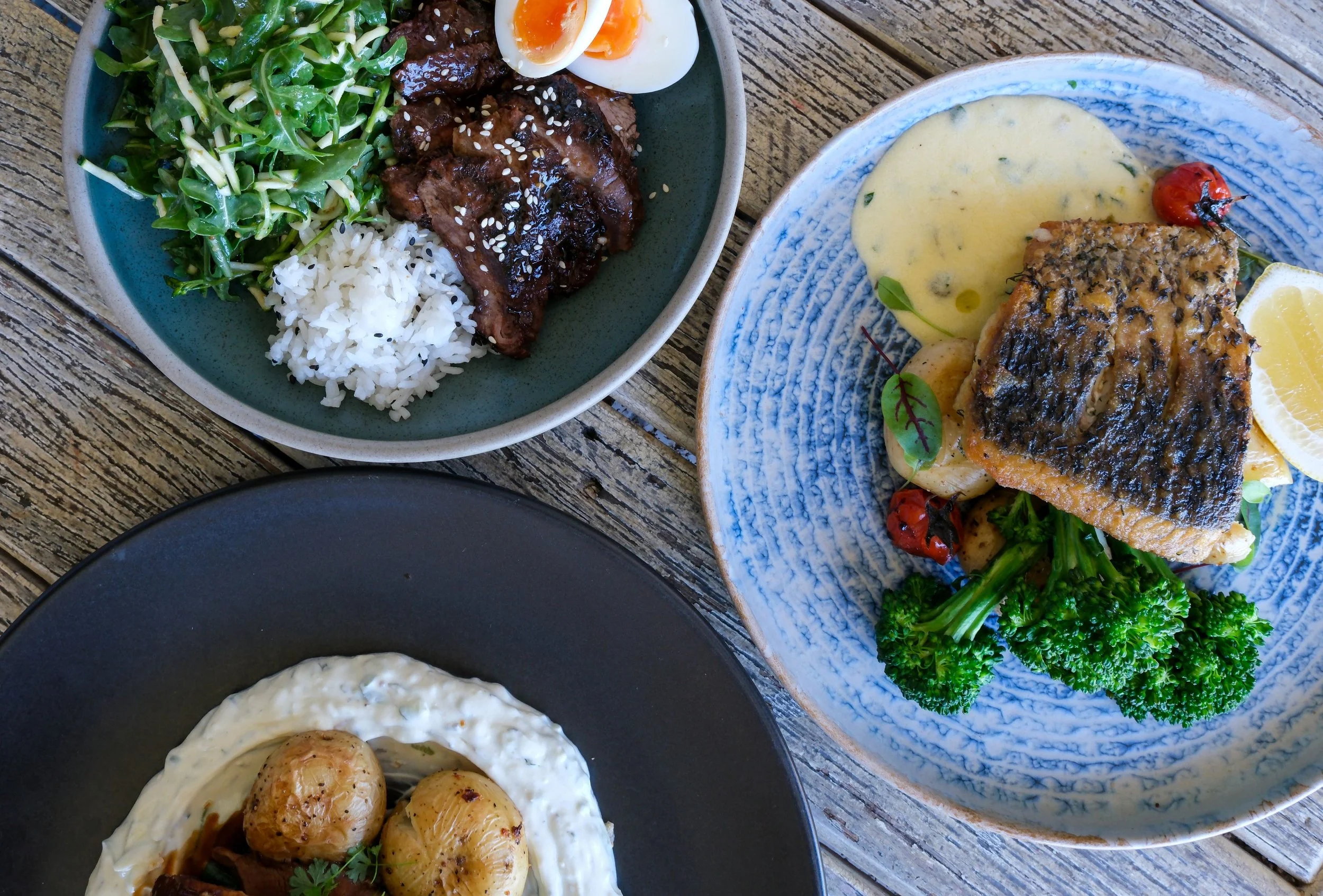 Various dishes on a wooden table, including a plate with rice, salad, grilled meat with sesame seeds, and boiled eggs; another plate with cooked fish, cherry tomatoes, lemon, broccoli, and a creamy sauce; and a black bowl with roasted potatoes in a white sauce.