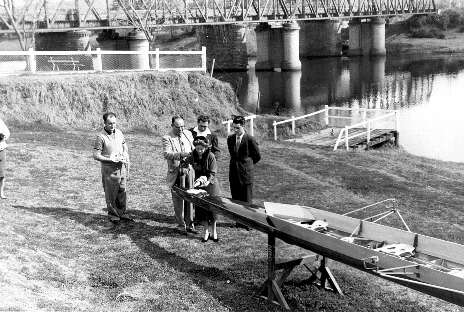 A black-and-white photo showing a group of five people standing near a model boat on a wooden stand outdoors, with a body of water, a bridge, and a grassy hillside in the background.