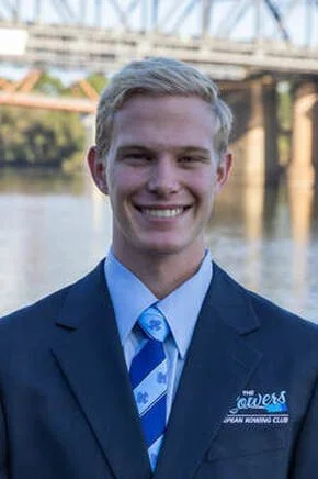 A young man in a suit standing outdoors near a body of water with a bridge in the background.