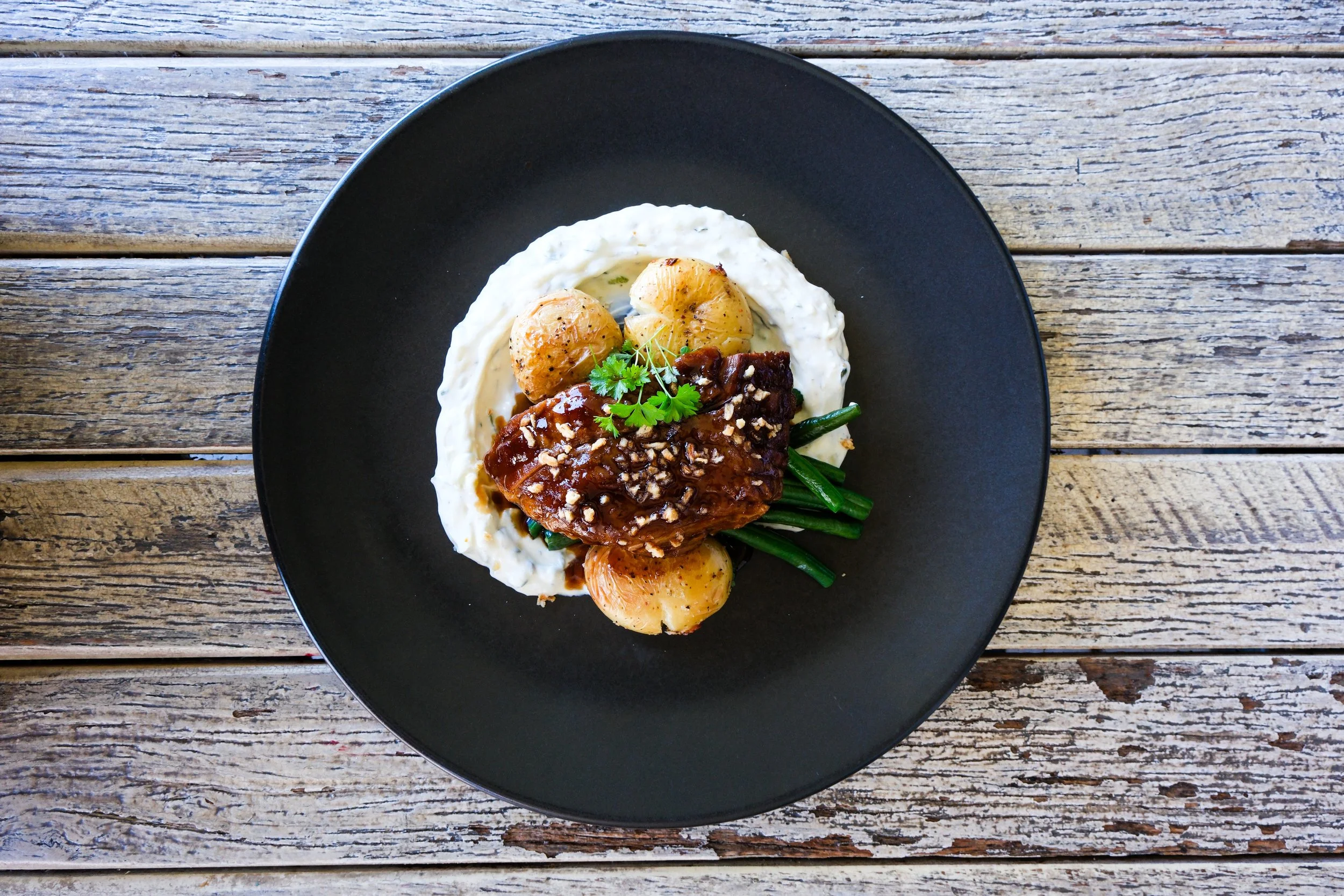 Black plate with a serving of meat topped with sauce and chopped nuts, boiled potatoes, green beans, and parsley garnish on a wooden table.