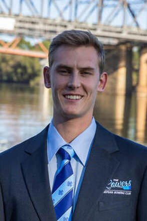 Young man in a suit smiling outdoors near a river with a bridge in the background.