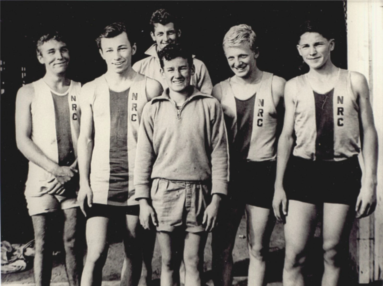 Black and white photo of five teenage boys and one young man in athletic clothing, standing outdoors.