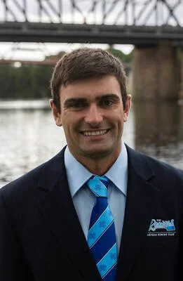 A man in a dark suit and blue tie smiling outdoors with a bridge and water in the background.