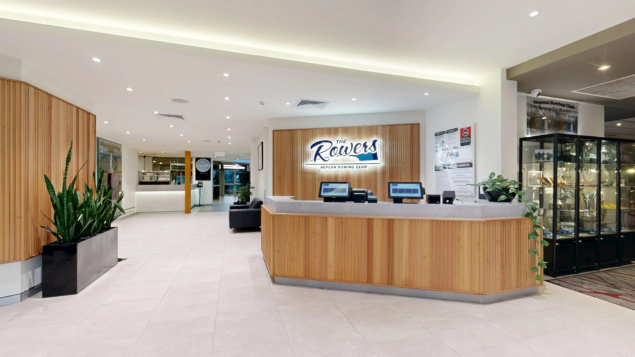 Reception area of The Rowers Nepean Rowing Club with a wooden front desk, two computer terminals, indoor plants, and display case with trophies.
