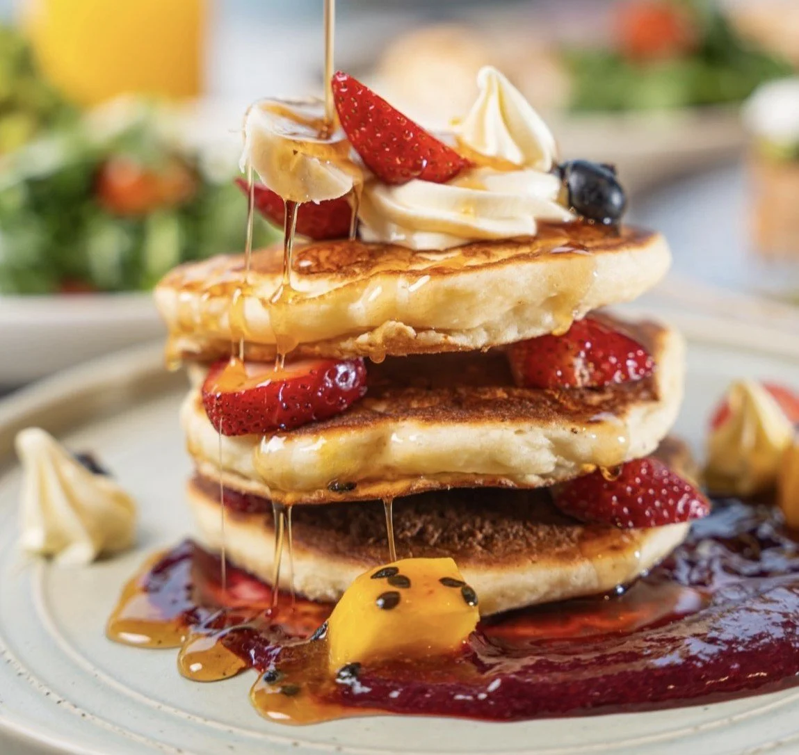 Stacked pancakes topped with strawberries, bananas, whipped cream, and syrup, with extra berries and syrup on the plate.