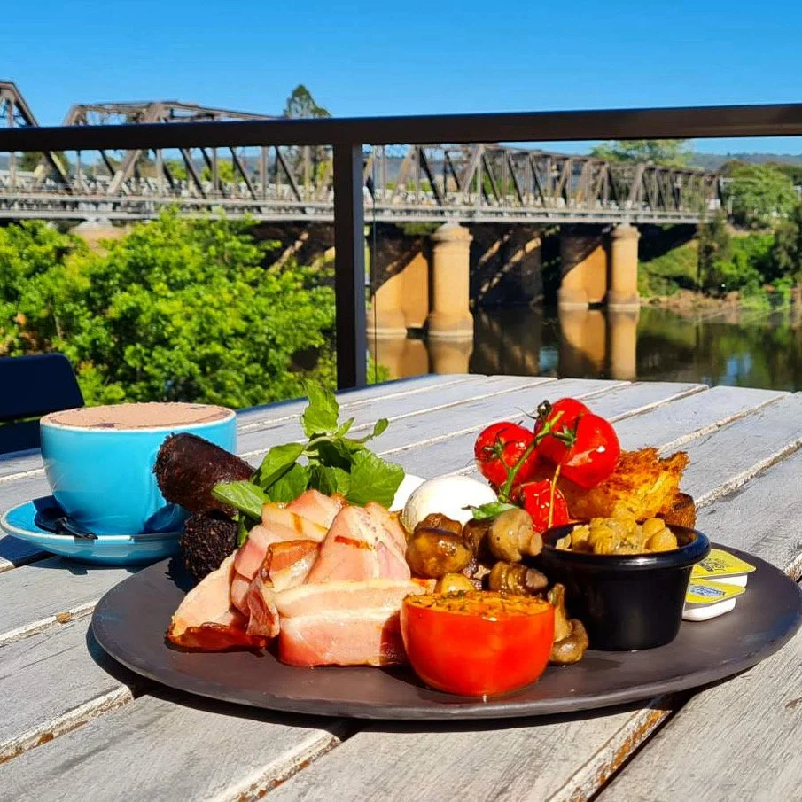 A breakfast tray with bacon, cherry tomatoes, mushrooms, an egg, toast, herbs, and a cup of hot chocolate or coffee on a wooden table outdoors with a bridge and river in the background.