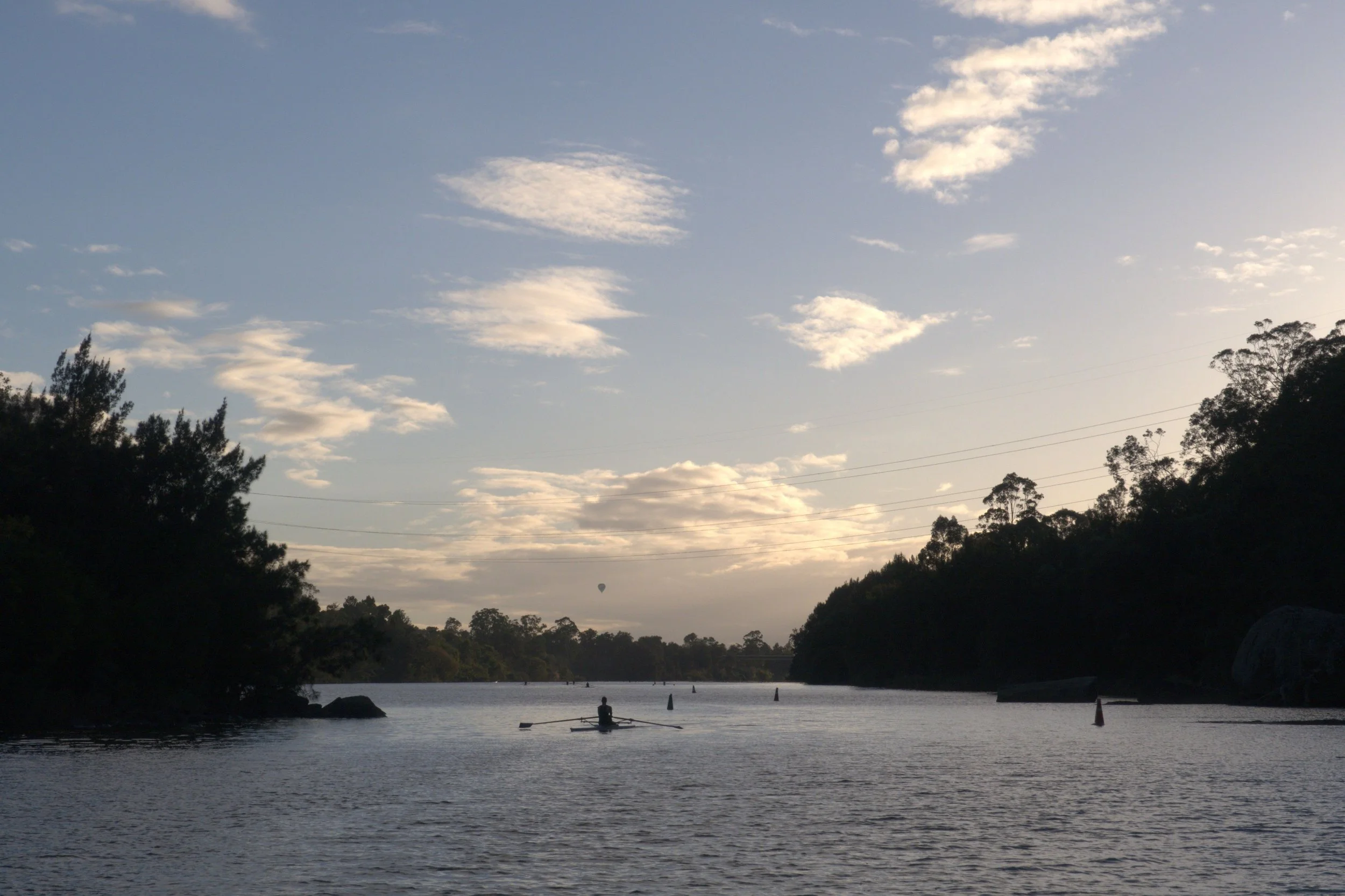 A calm river scene during sunset with a person in a small boat, trees along the riverbanks, and a hot air balloon visible in the distance against a partly cloudy sky.