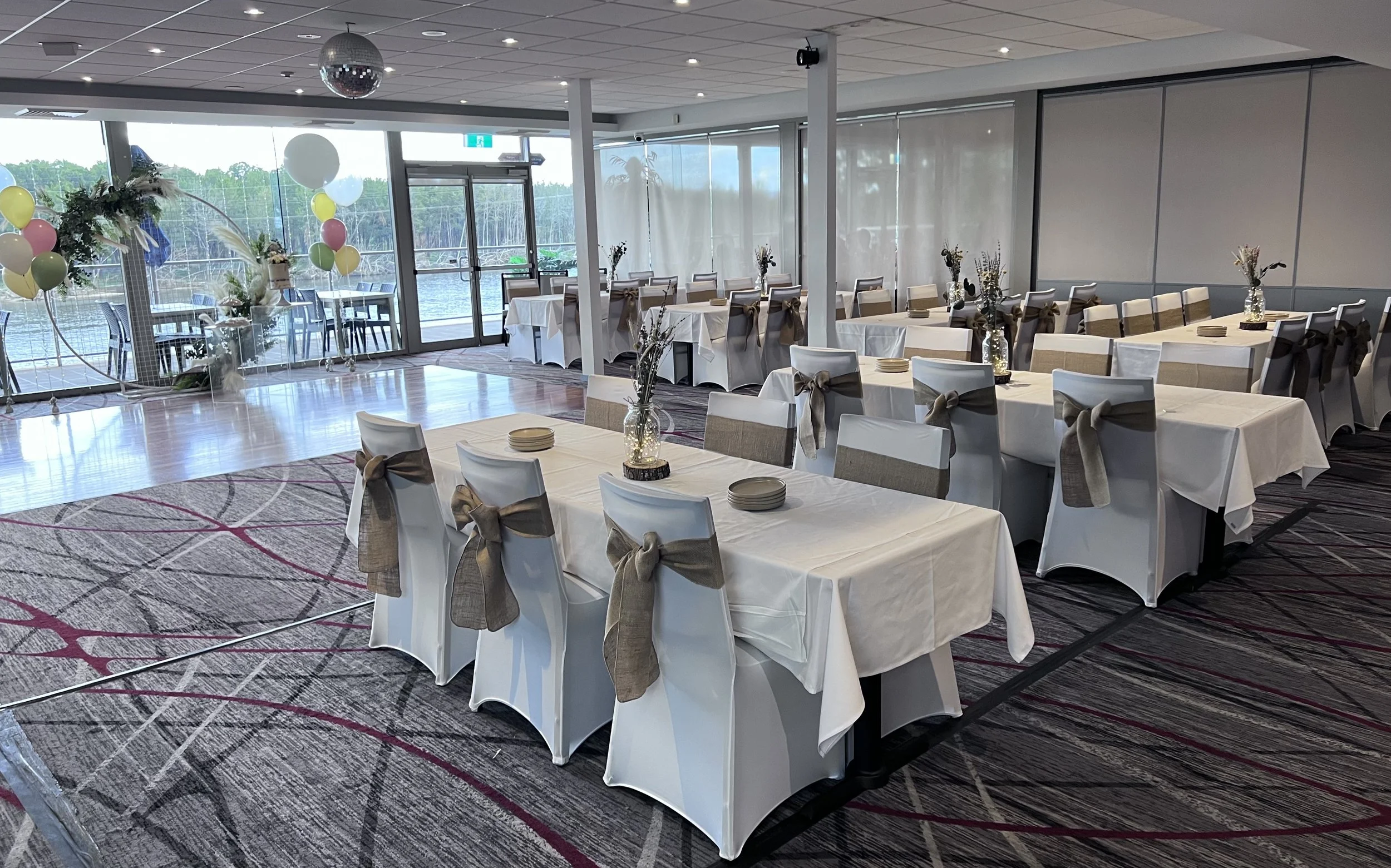 A decorated banquet hall with tables covered in white tablecloths, each with a vase of flowers and brown bows on the chairs, set for a celebration near a large window overlooking a body of water, with colorful balloons and balloons arches in the corner.
