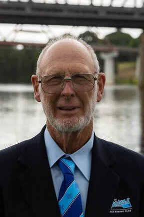 Close-up of an older man with glasses, a beard, and a receding hairline, wearing a suit and tie, standing outdoors near a body of water with trees and a bridge in the background.