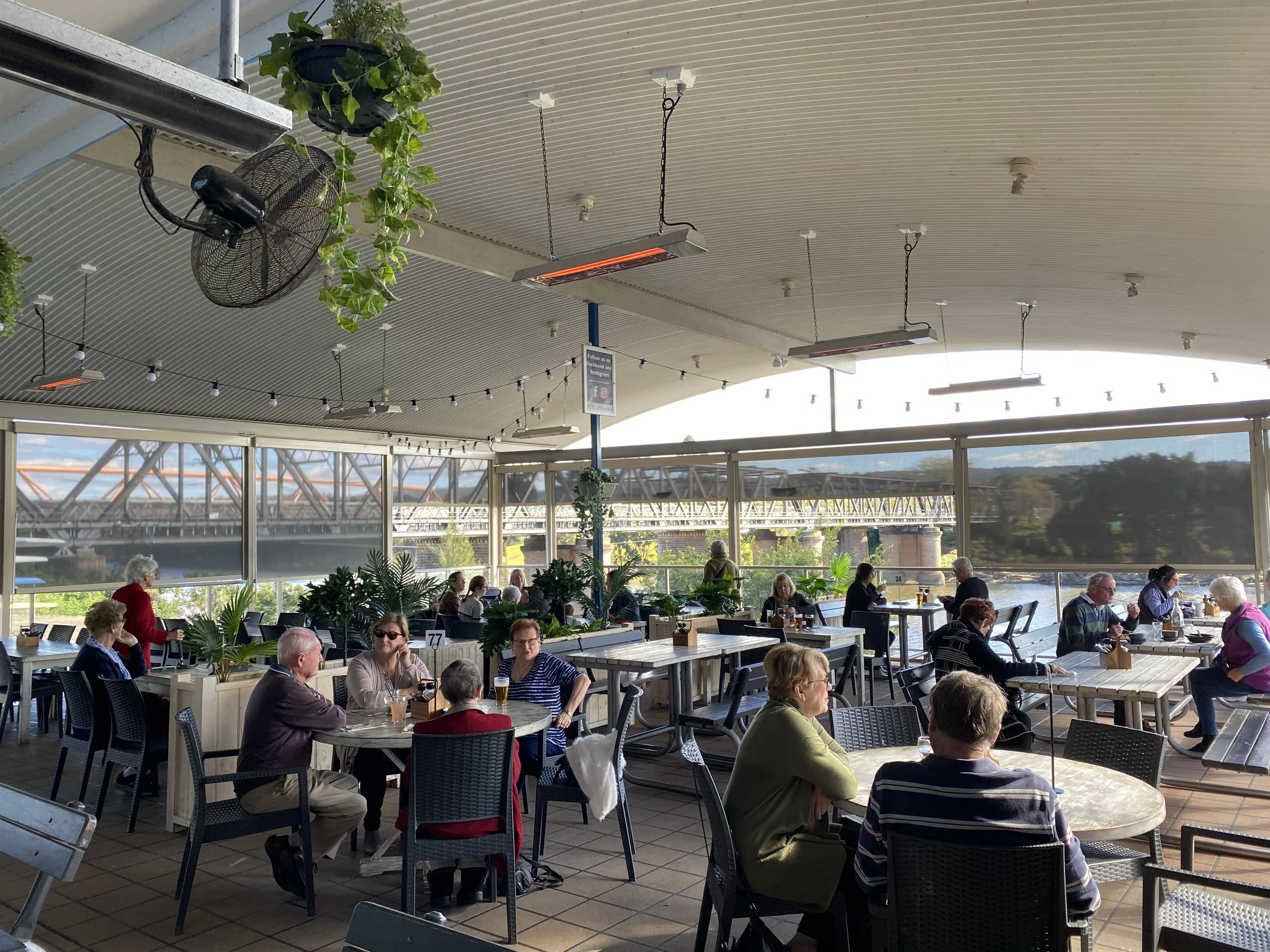 People dining in an outdoor restaurant or cafe under a curved roof with hanging heater lamps and string lights, overlooking a bridge and river.